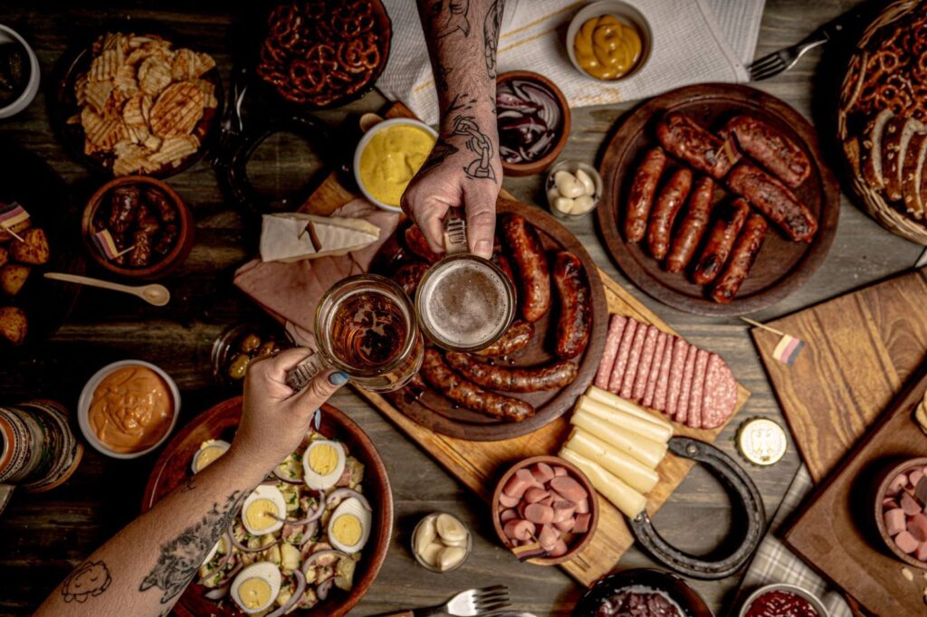 Overhead shot of a rustic meal featuring sausages, snacks, and beer on a wooden table.